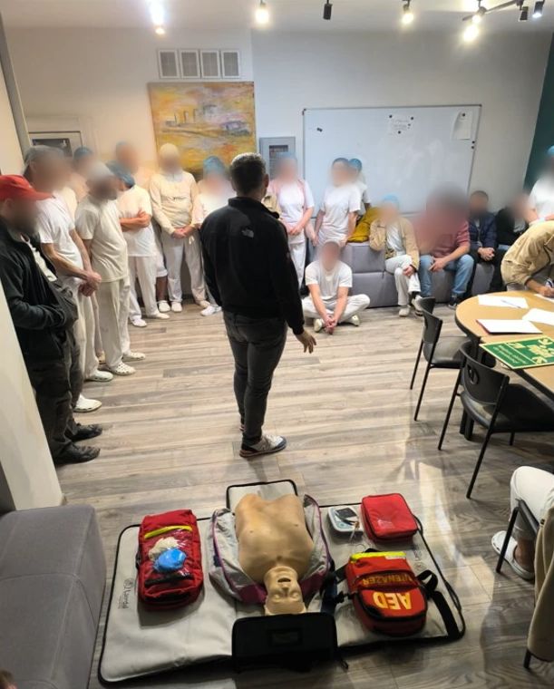 Sala szkoleniowa: na pierwszym planie fantom do resuscytacji między torbą z defibrylatorem AED a apteczką, dalej prowadzący stoi przed kilkunastoosobową grupą uczestników.