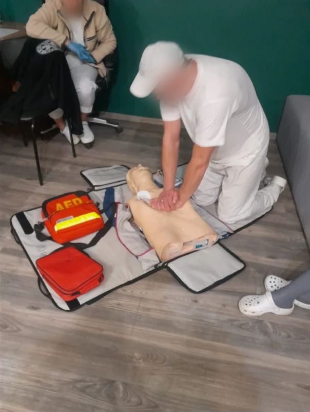 Uczestnik w białym ubraniu klęczy przy fantomie i oburącz uciska jego klatkę piersiową, obok na macie leżą torba z defibrylatorem AED i czerwona apteczka.